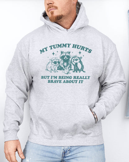 Man wearing an ash gray hoodie with a retro raccoon gang graphic and the quote 'My Tummy Hurts But I'm Being Really Brave About It'.