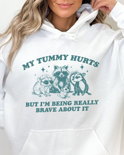 Close-up of white hoodie with a retro raccoon gang graphic and the quote 'My Tummy Hurts But I'm Being Really Brave About It'.