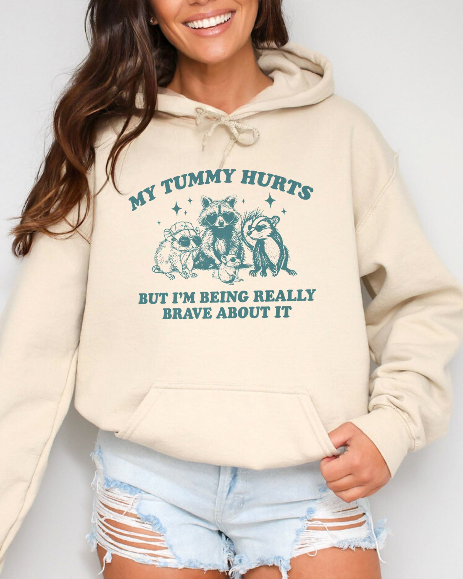 Woman wearing a sand beige hoodie with a retro raccoon gang graphic and the quote 'My Tummy Hurts But I'm Being Really Brave About It'.