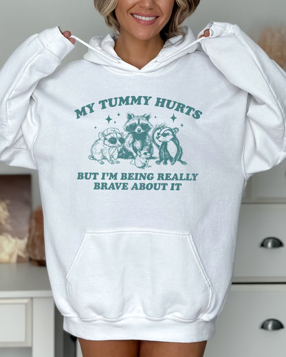 Woman wearing a white hoodie featuring a funny retro raccoon gang graphic and the quote 'My Tummy Hurts But I'm Being Really Brave About It'.