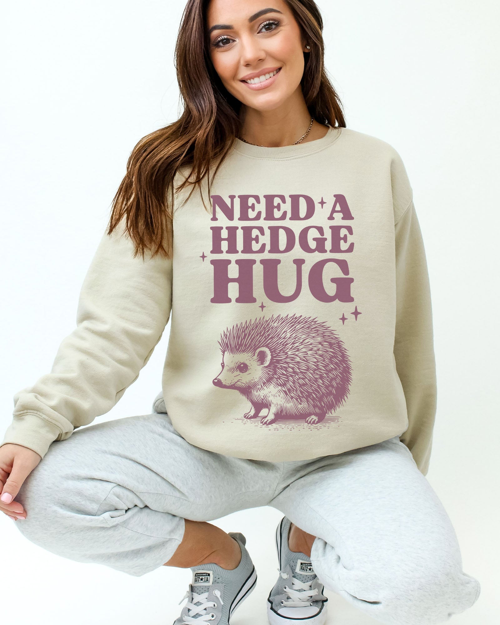 Model wearing sand color sweatshirt with 'Need A Hedge Hug' graphic design