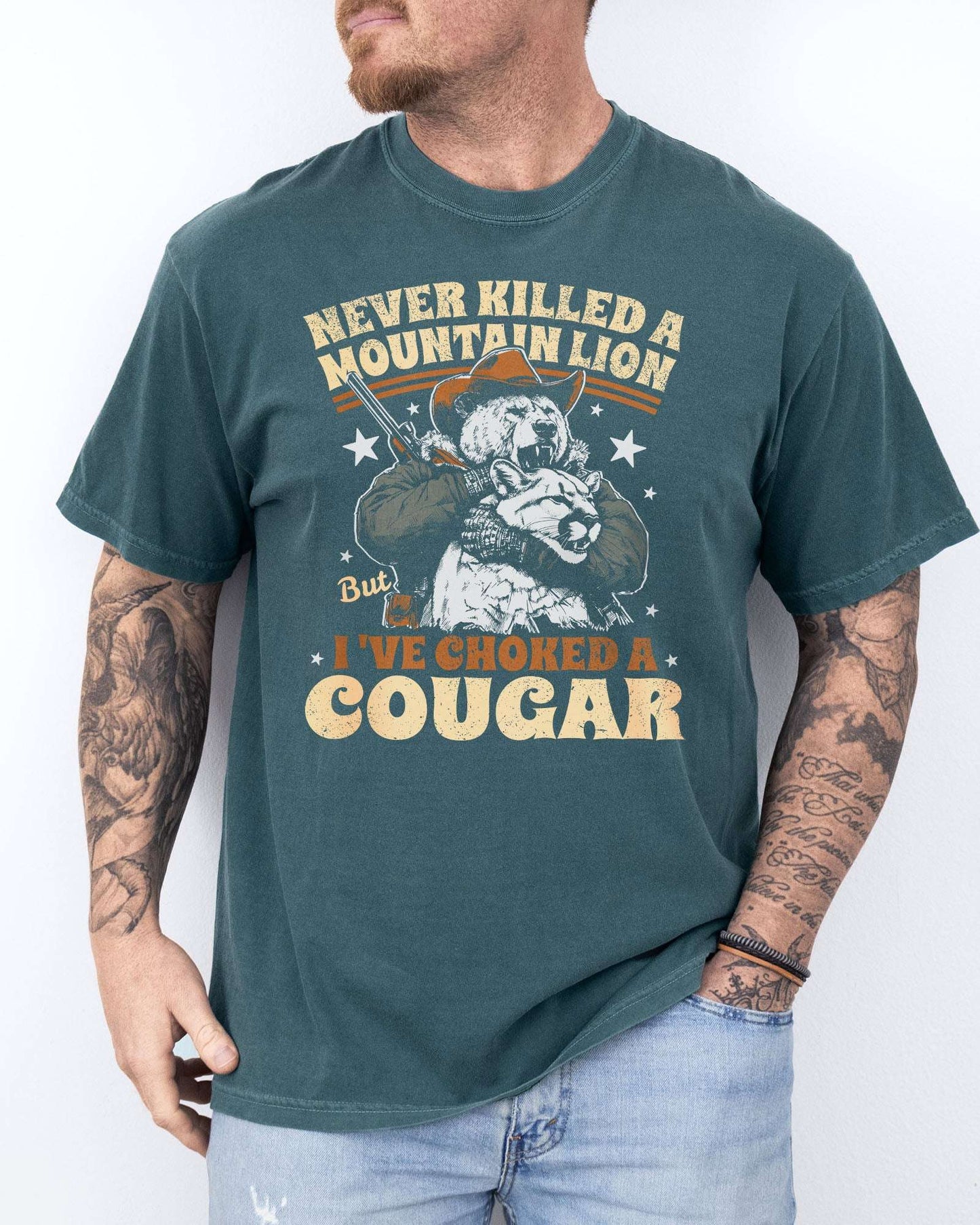 Man wearing a blue spruce "Never Killed a Mountain Lion But I've Choked a Cougar" shirt with a funny retro graphic of a bear choking a cougar.