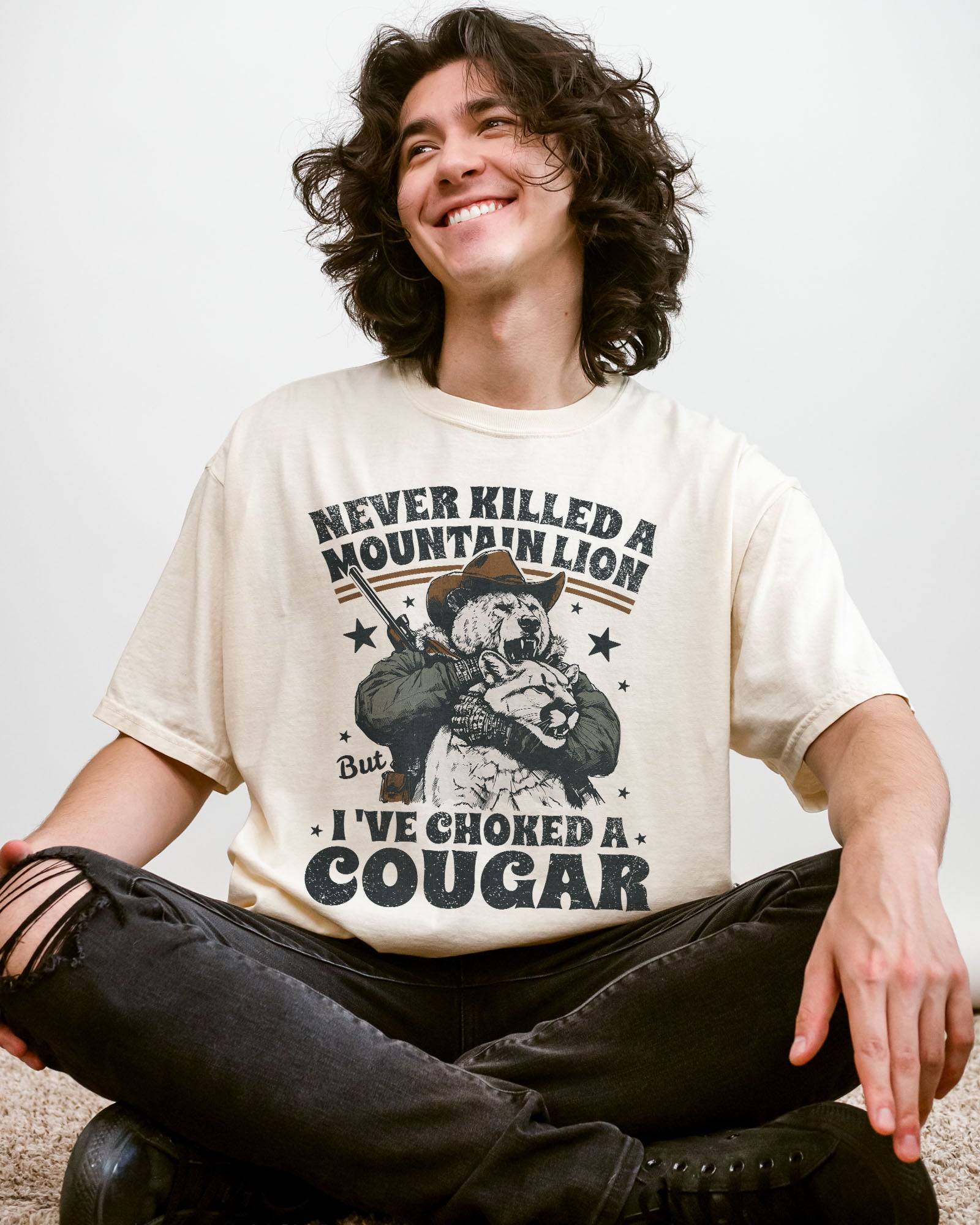 Man wearing an ivory "Never Killed a Mountain Lion But I've Choked a Cougar" shirt with a funny retro graphic of a bear choking a cougar.