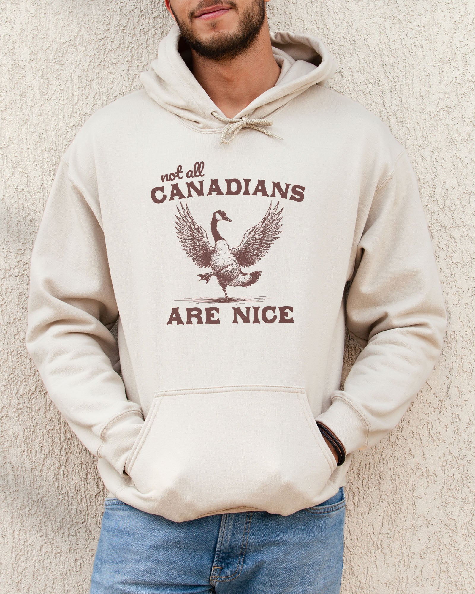 Man wearing sand-colored 'Not All Canadians Are Nice' hoodie featuring a funny canadian goose retro graphic.