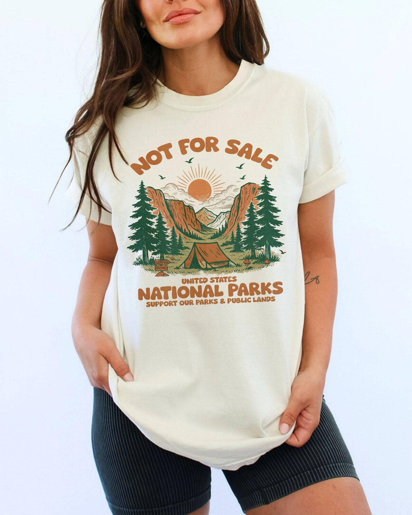 Woman wearing an ivory 'Not For Sale - United States National Parks' shirt with a retro graphic of national park woods and a tent.
