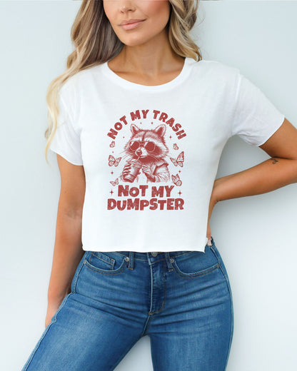 Woman wearing a white 'Not My Trash Not My Dumpster' crop top with a funny retro raccoon drinking coffee graphic.