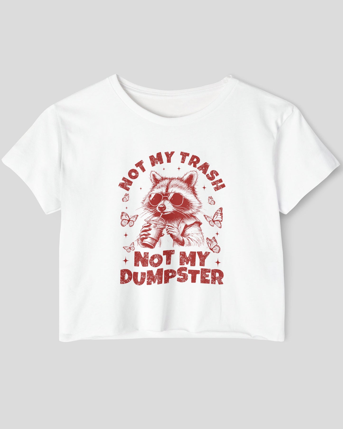 Flat lay of a white 'Not My Trash Not My Dumpster' crop top featuring a funny retro raccoon drinking coffee graphic.