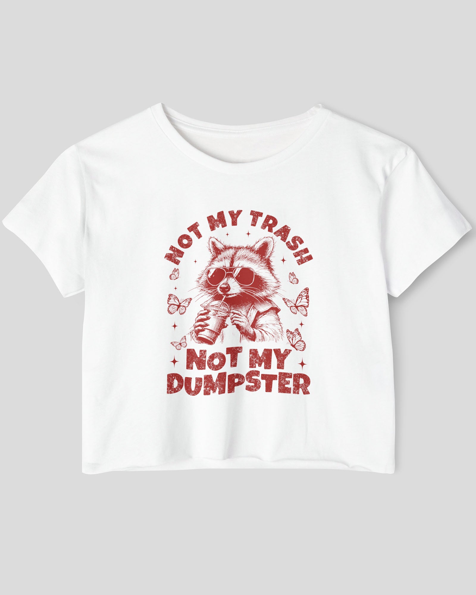 Flat lay of a white 'Not My Trash Not My Dumpster' crop top featuring a funny retro raccoon drinking coffee graphic.