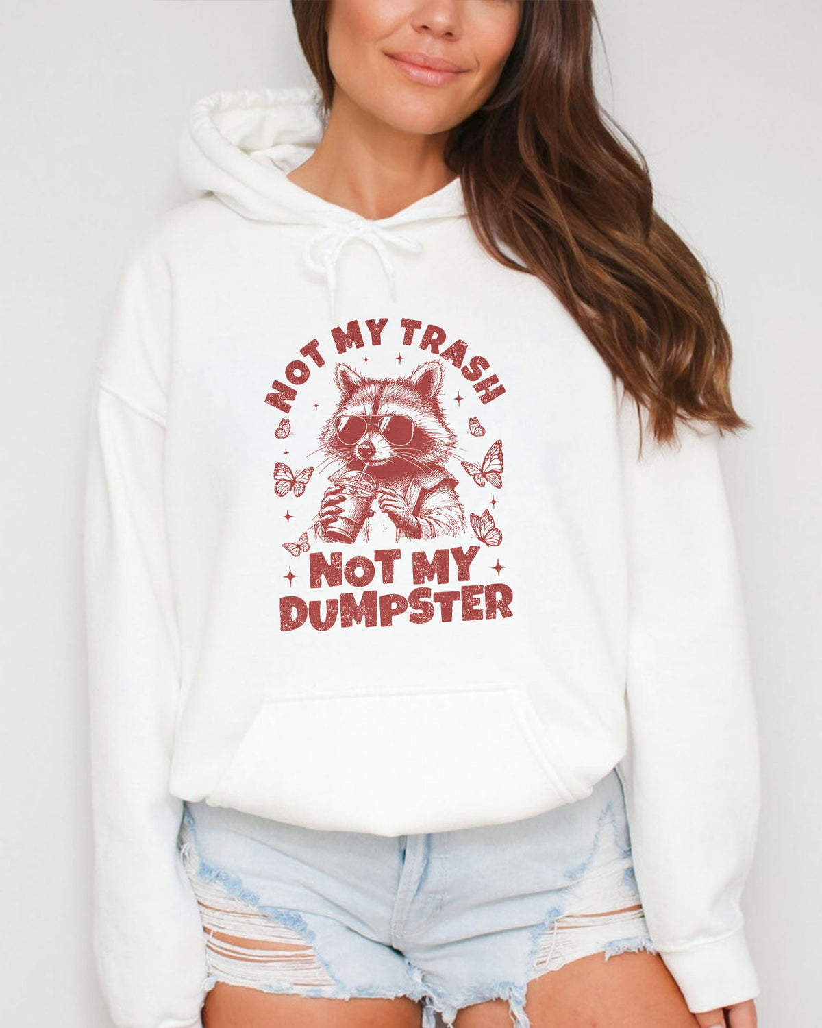 Woman wearing a white 'Not My Trash, Not My Dumpster' hoodie with a funny retro raccoon drinking coffee graphic.
