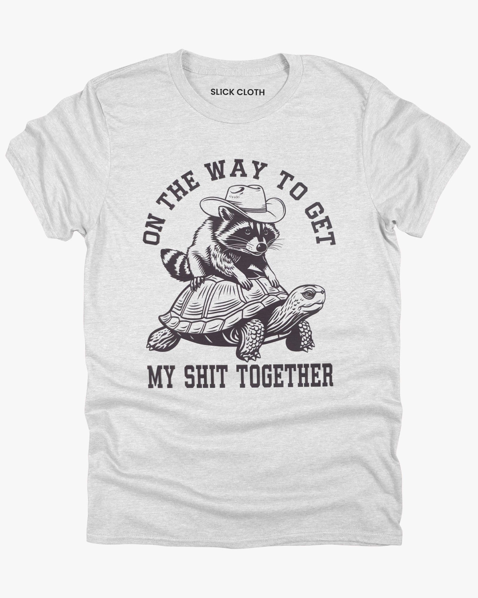 Flat lay ash gray 'On The Way to Get My Shit Together' t-shirt featuring a funny retro raccoon riding a slow-moving turtle graphic.