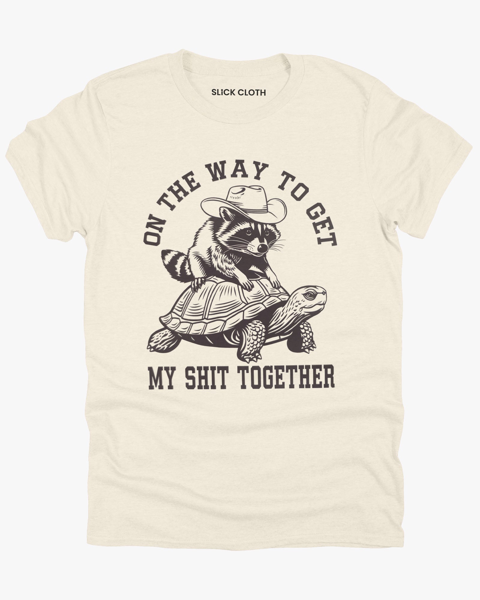 Flat lay natural color 'On The Way to Get My Shit Together' shirt featuring a funny retro raccoon riding a slow-moving turtle graphic.