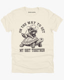 Flat lay natural color 'On The Way to Get My Shit Together' shirt featuring a funny retro raccoon riding a slow-moving turtle graphic.