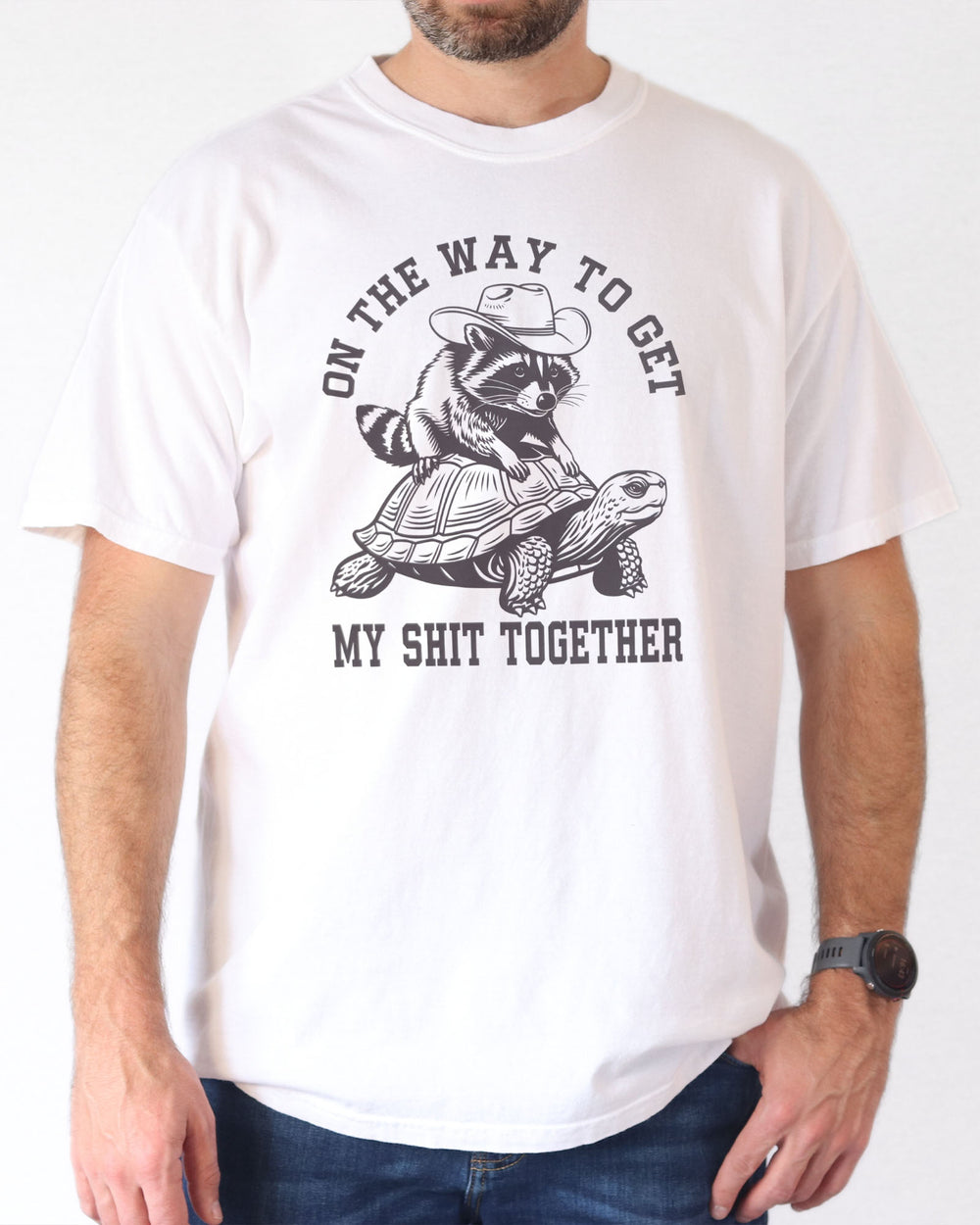 Man wearing a white 'On The Way to Get My Shit Together' t-shirt featuring a funny retro raccoon riding a slow-moving turtle graphic.