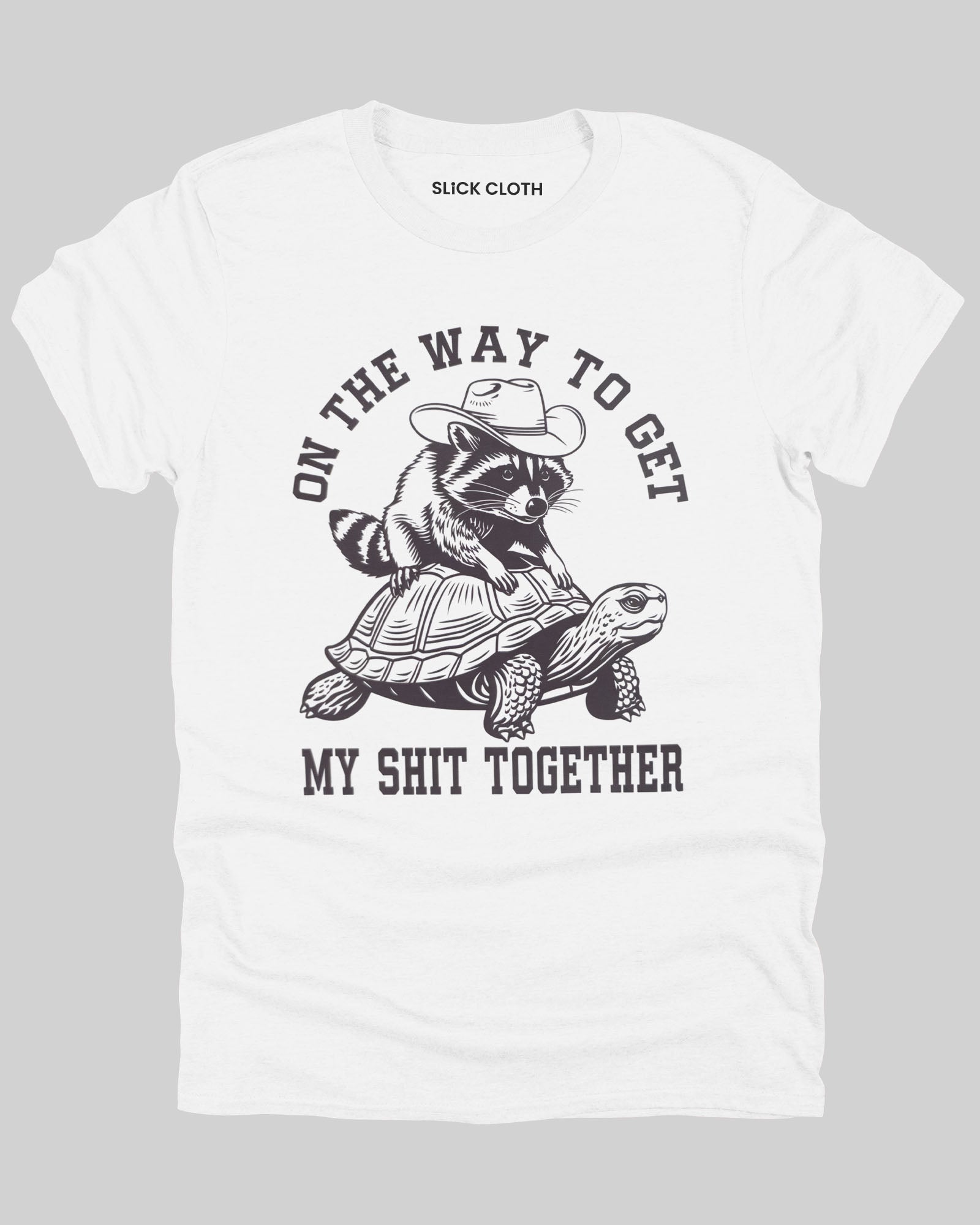 Flat lay white color 'On The Way to Get My Shit Together' t-shirt featuring a funny retro raccoon riding a slow-moving turtle graphic.
