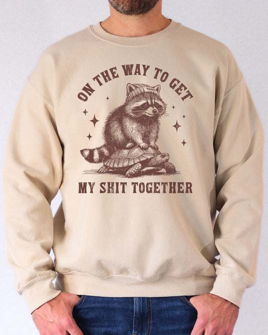 Man wearing a sand beige sweatshirt featuring a funny retro graphic of a raccoon riding a tortoise with the quote 'On The Way To Get My Shit Together'.