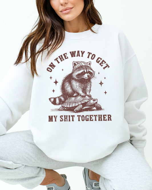 Woman wearing a white sweatshirt featuring a funny retro graphic of a raccoon riding a tortoise with the quote 'On The Way To Get My Shit Together'.