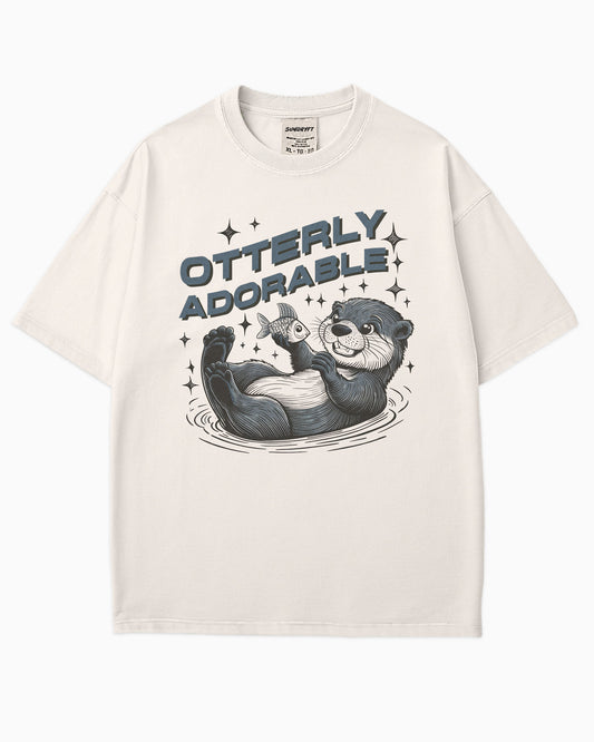 Flat lay ivory 'Otterly Adorable' shirt with a retro graphic of a happy otter holding a fish.