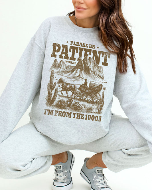 Woman wearing an ash gray sweatshirt featuring a retro western scene with a horse-drawn carriage in the desert and the quote 'Please Be Patient With Me, I'm From the 1900s'.