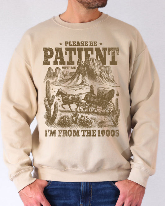 Man wearing a sand beige sweatshirt featuring a retro western scene with a horse-drawn carriage in the desert and the quote 'Please Be Patient With Me, I'm From the 1900s'.