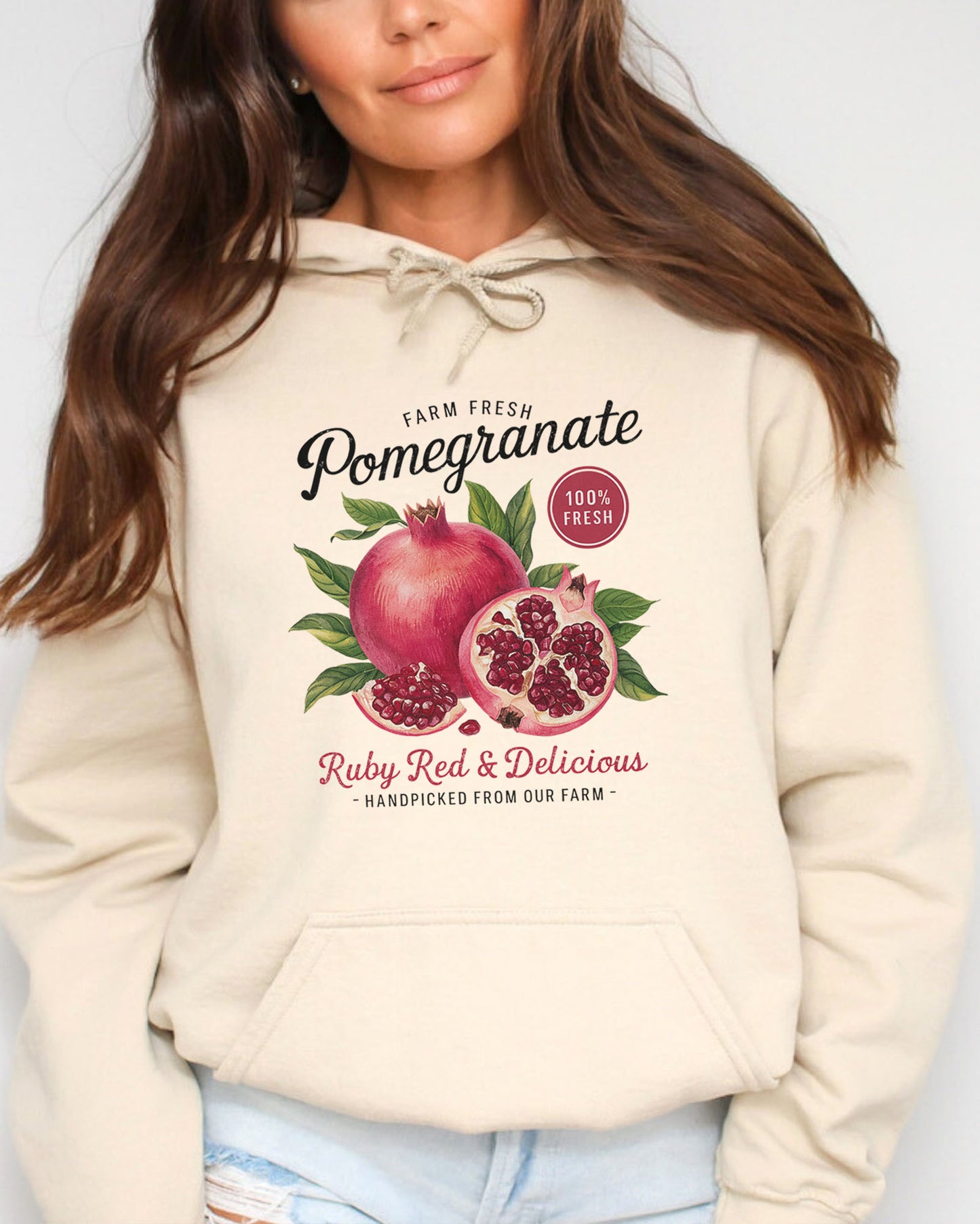 Woman wearing a sand beige hoodie featuring a vintage-style Pomegranate fruit label graphic with the text 'Pomegranate - Ruby Red & Delicious'.