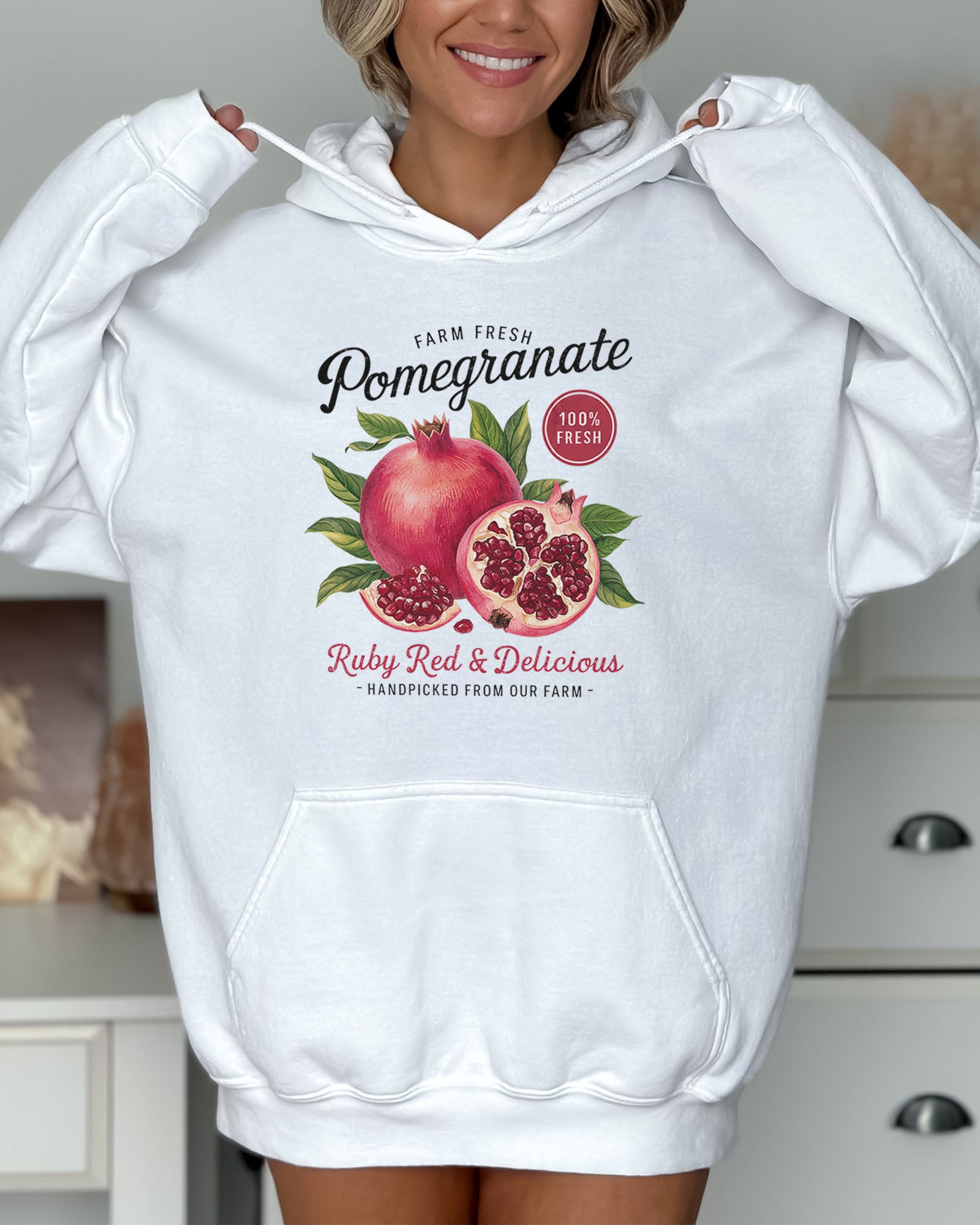 Woman wearing a white hoodie featuring a vintage-style Pomegranate fruit label graphic with the text 'Pomegranate - Ruby Red & Delicious'.