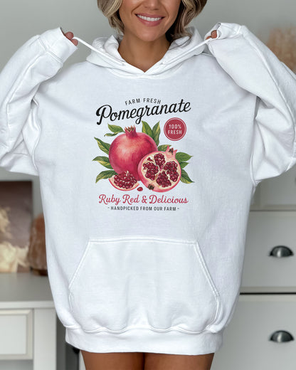 Woman wearing a white hoodie featuring a vintage-style Pomegranate fruit label graphic with the text 'Pomegranate - Ruby Red & Delicious'.