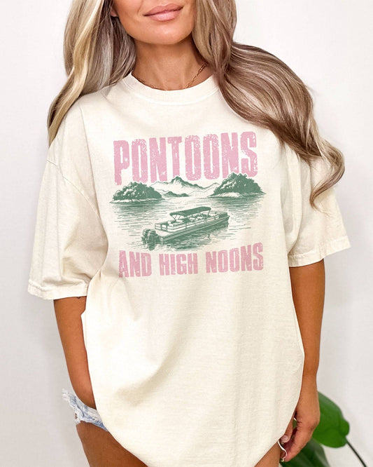 Woman wearing "Pontoons And High Noons" shirt in ivory color with a retro graphic of a pontoon on a lake.
