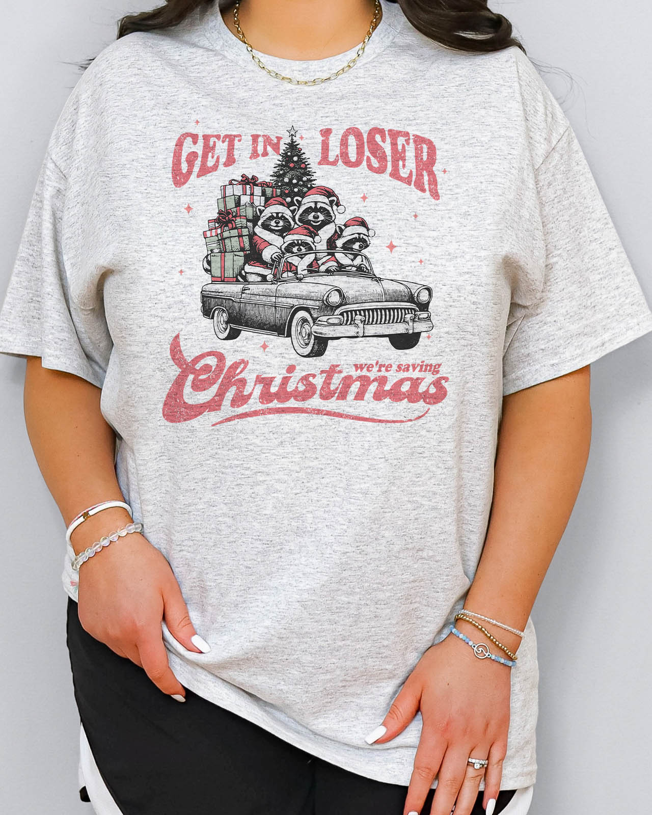 Woman wearing "Get In Loser We're Saving Christmas" shirt in ash grey color with a retro funny raccoon XMAS graphic