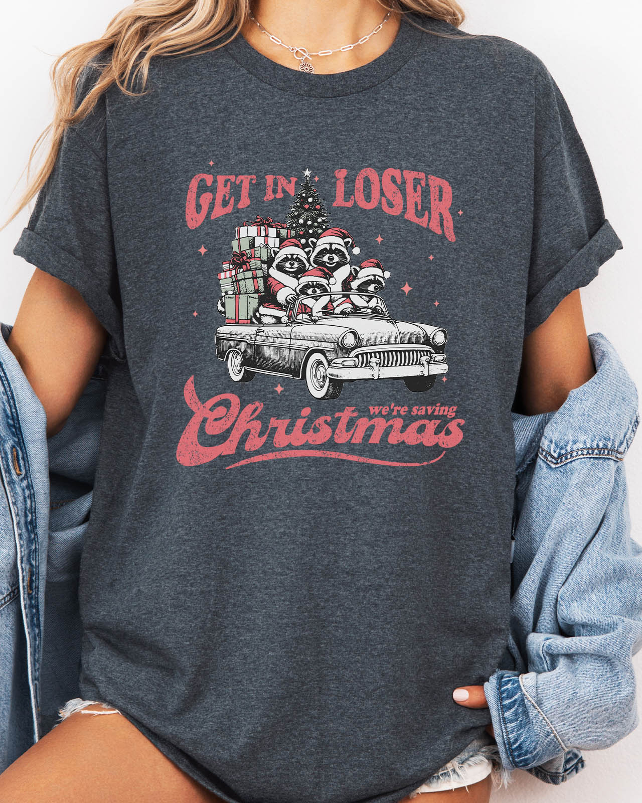 Woman wearing "Get In Loser We're Saving Christmas" shirt in dark heather color with a retro funny raccoon XMAS graphic
