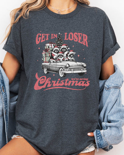 Woman wearing "Get In Loser We're Saving Christmas" shirt in dark heather color with a retro funny raccoon XMAS graphic