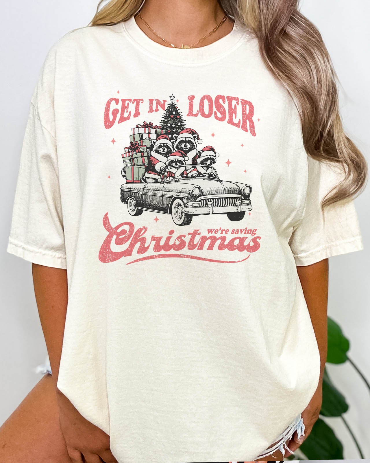 Woman wearing "Get In Loser We're Saving Christmas" shirt in natural beige color with a retro funny raccoon XMAS graphic