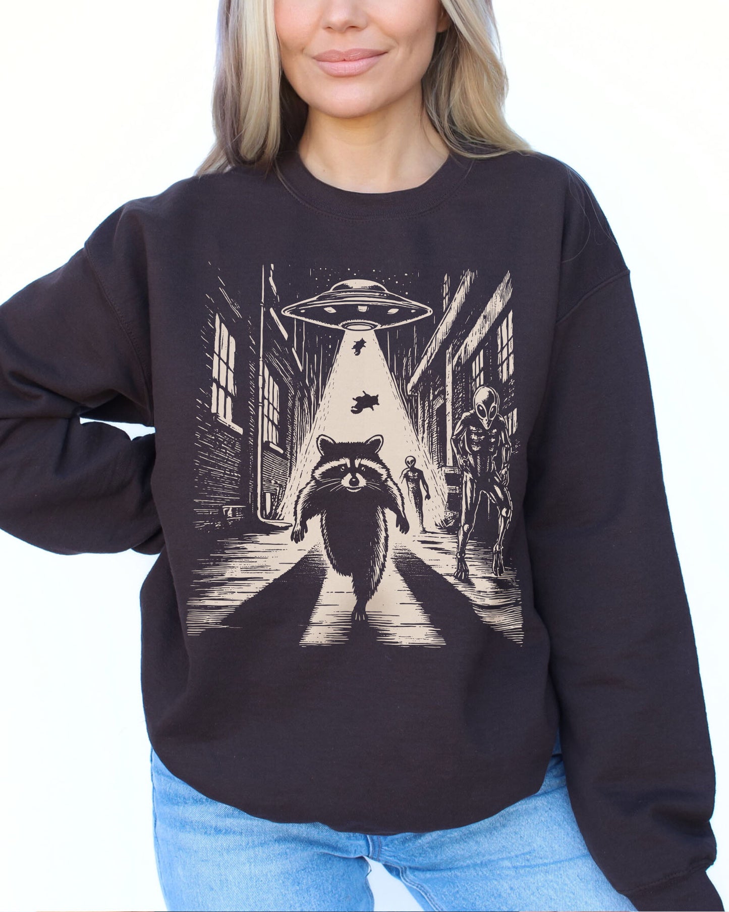 Woman wearing a dark chocolate sweatshirt with a retro graphic of a raccoon running from aliens.