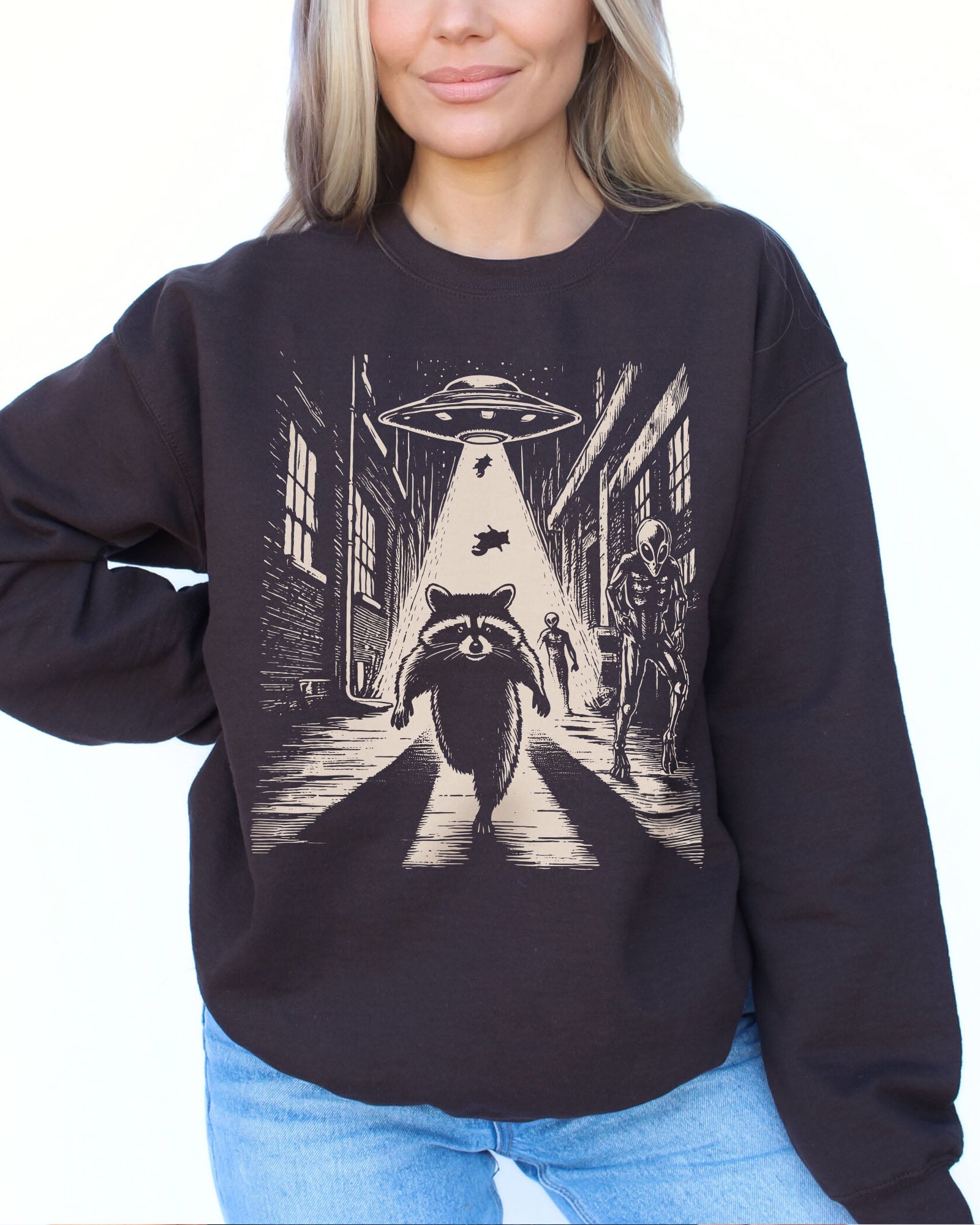 Woman wearing a dark chocolate sweatshirt with a retro graphic of a raccoon running from aliens.