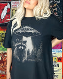 Woman wearing a black 'Raccoon UFO' t-shirt featuring a funny graphic of a raccoon escaping a UFO abduction.
