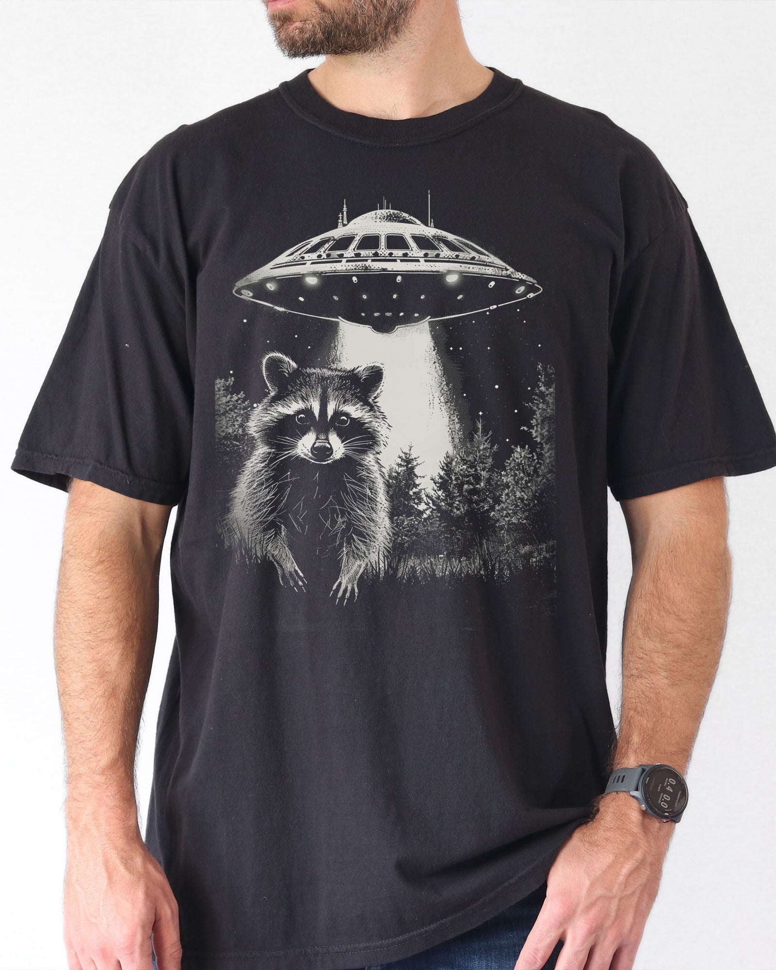 Man wearing a black 'Raccoon UFO' t-shirt featuring a funny graphic of a raccoon escaping a UFO abduction.