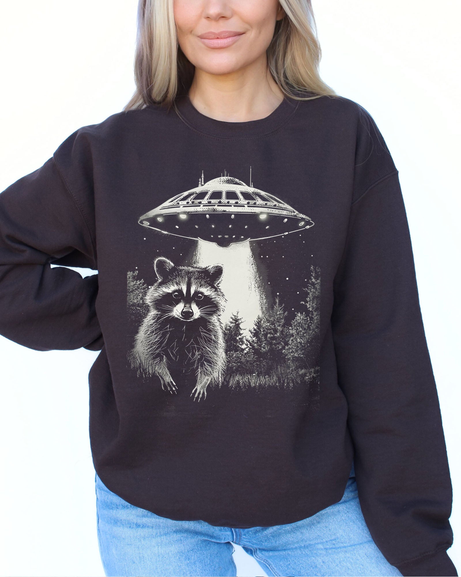 Model wearing dark chocolate color sweatshirt with 'Raccoon Escaping UFO Abduction' graphic design