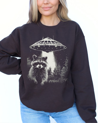 Model wearing dark chocolate color sweatshirt with 'Raccoon Escaping UFO Abduction' graphic design