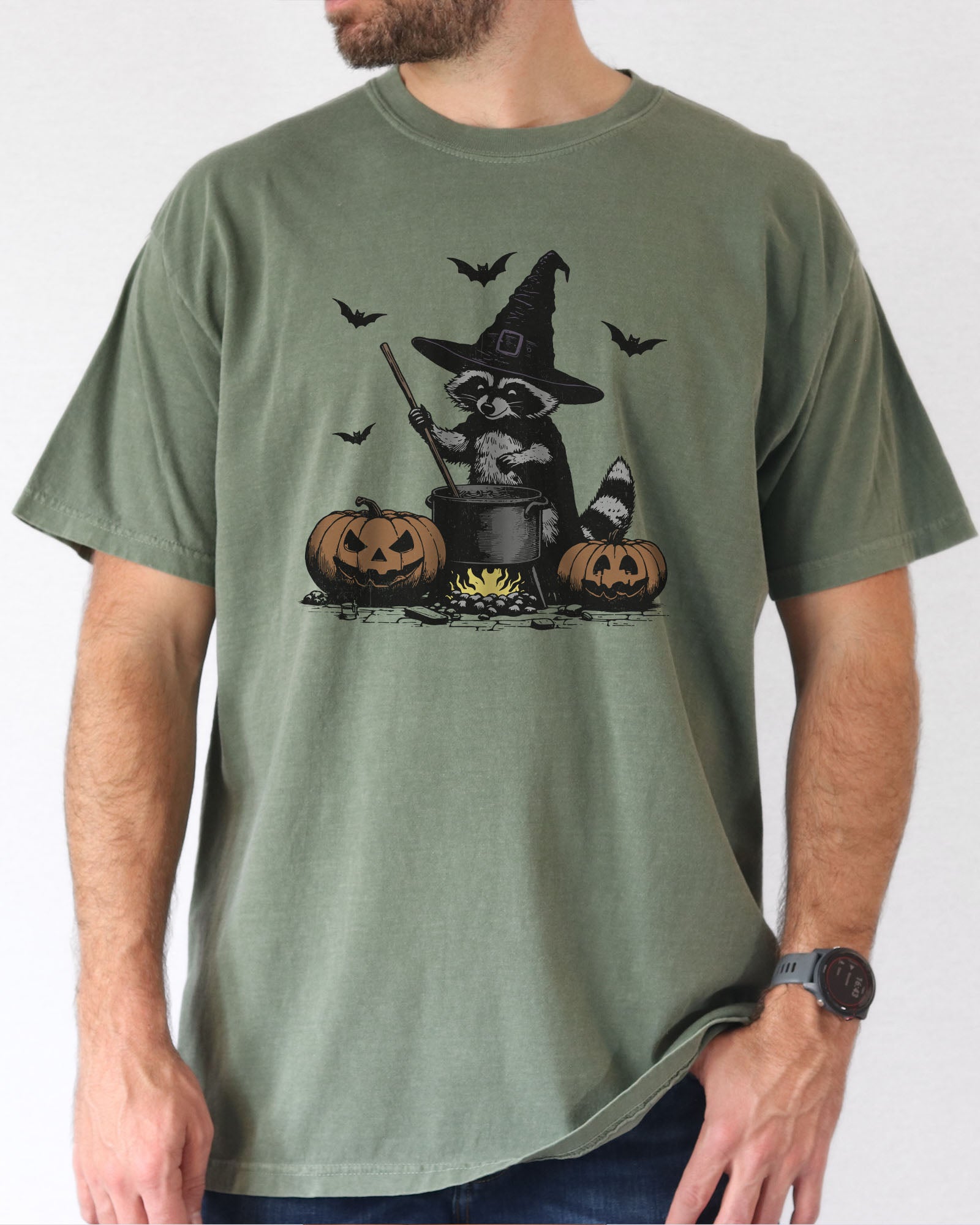 Model wearing moss color shirt with 'Halloween Raccoon Witch' graphic design
