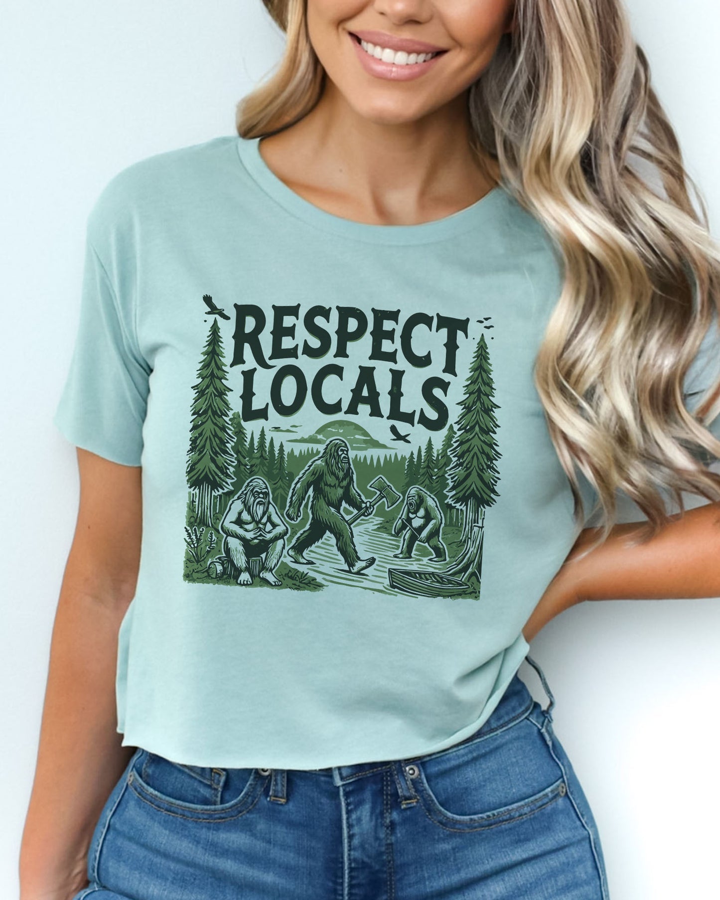 Woman wearing stonewash green 'Respect Locals' crop top featuring a funny retro Bigfoot walking through the forest graphic.