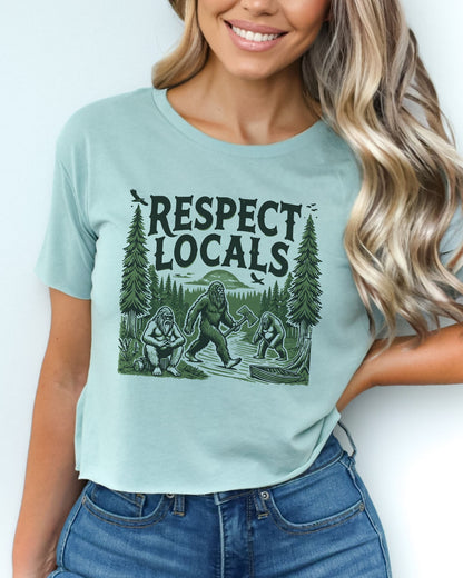 Woman wearing stonewash green 'Respect Locals' crop top featuring a funny retro Bigfoot walking through the forest graphic.