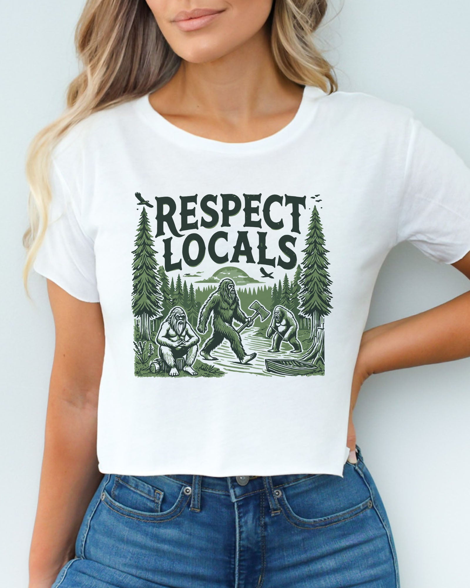 Woman wearing white 'Respect Locals' crop top featuring a funny retro Bigfoot walking through the forest graphic.