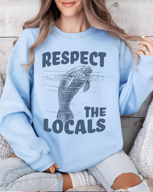 Woman wearing a light blue sweatshirt with a retro manatee graphic and the text “Respect the Locals.”