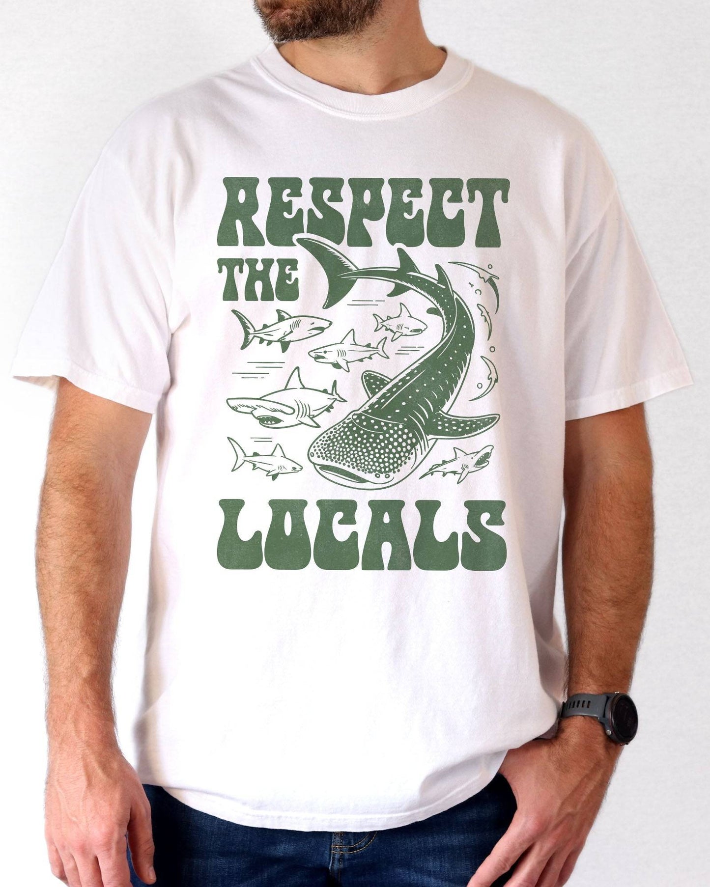 Man wearing a white-colored ‘Respect The Locals’ shirt with a retro graphic of whales, sharks, and fish.