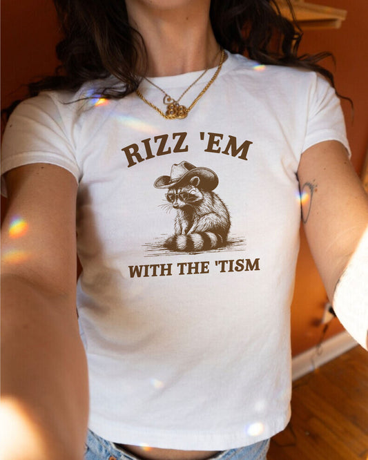 Woman wearing white color baby tee with the quote 'Rizz 'Em WIth The Tism' and a funny retro owboy raccoon graphic.