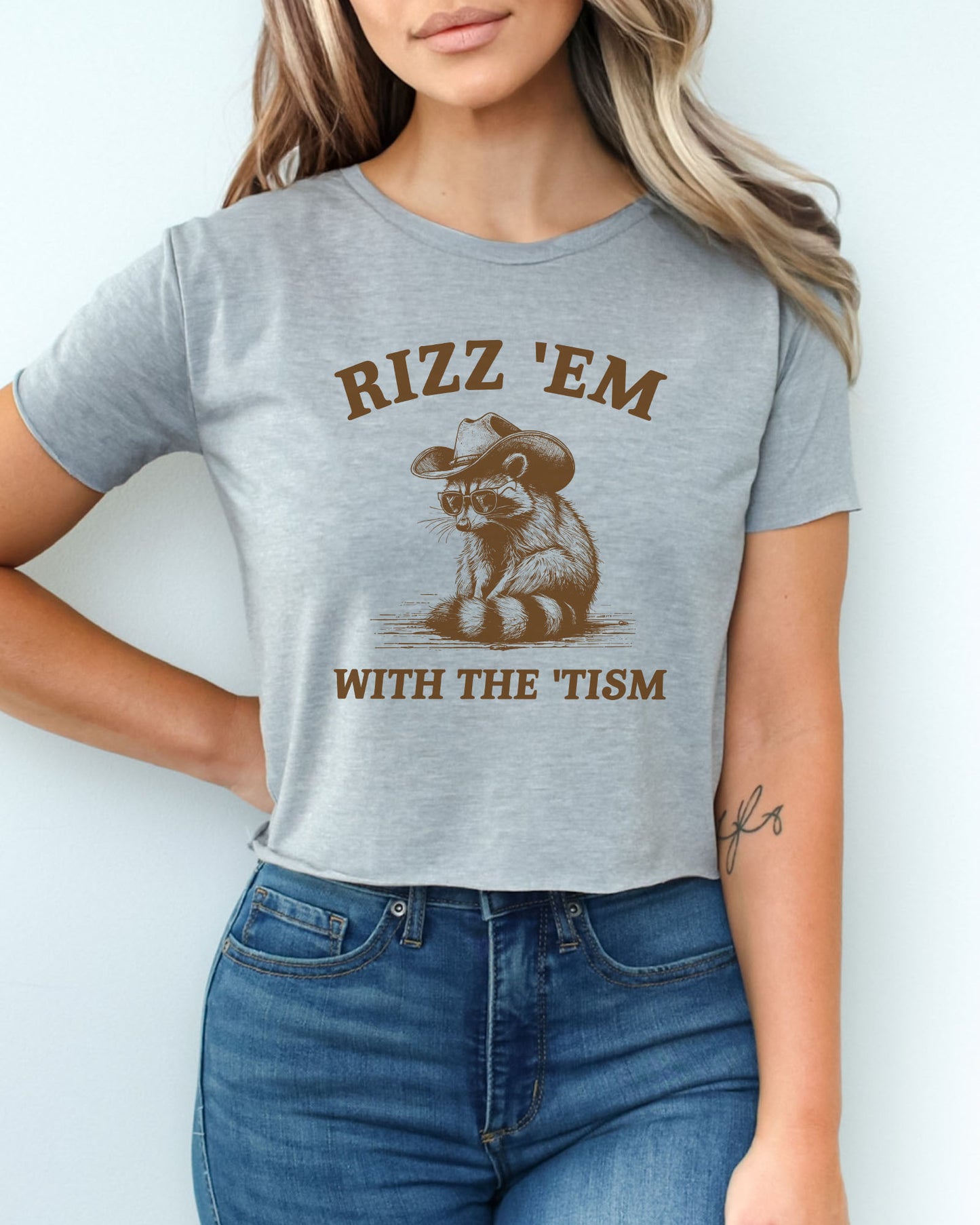 Woman wearing a heather gray 'Rizz Em With The Tism' crop top featuring a funny retro cowboy raccoon graphic.