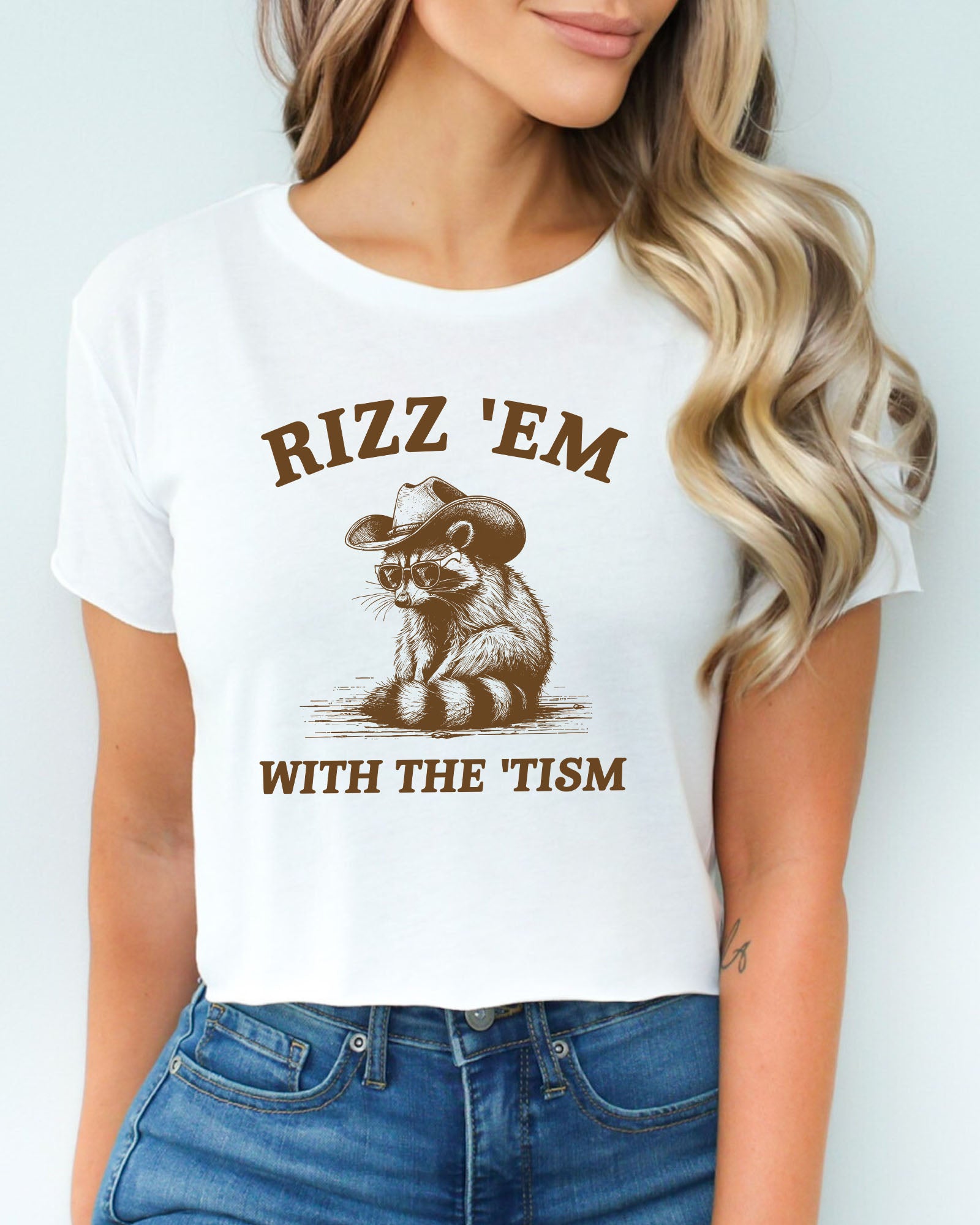 Woman wearing a white 'Rizz Em With The Tism' crop top featuring a funny retro cowboy raccoon graphic.