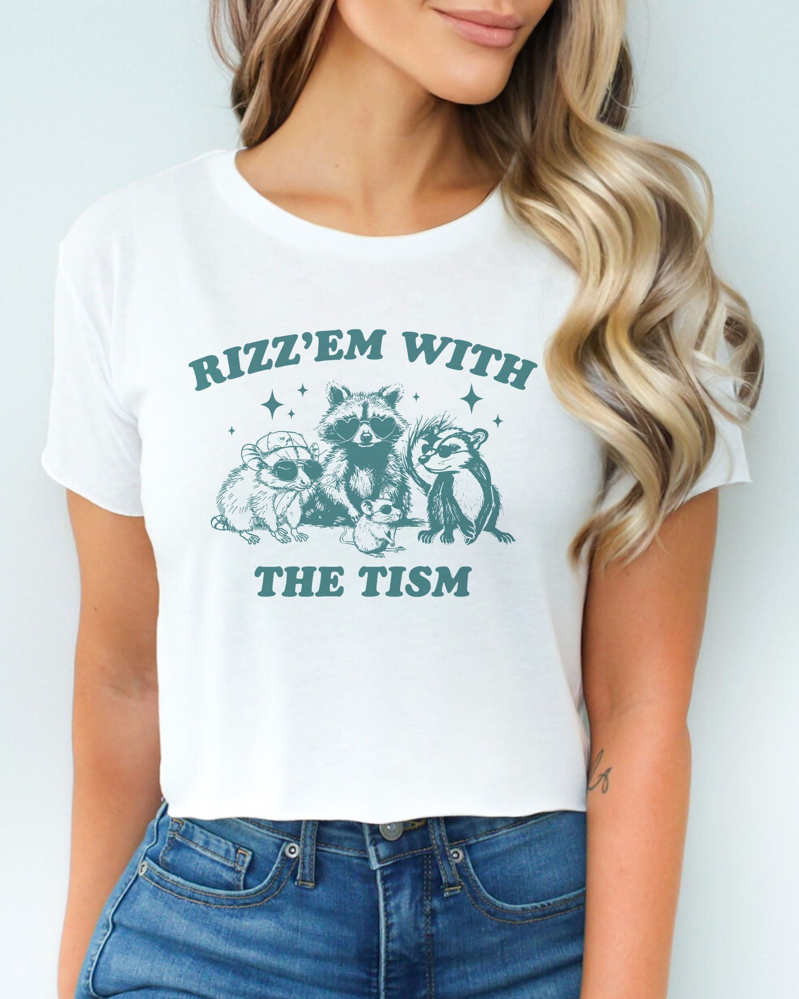 Woman wearing a white 'Rizz 'Em With The Tism' crop top featuring a funny retro raccoon gang graphic. Trendy and humorous women's graphic crop tee.