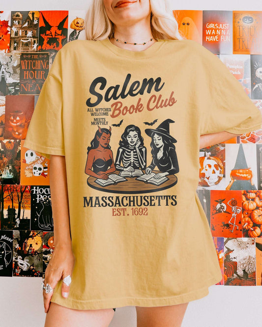 Woman wearing a mustard-colored 'Salem Book Club Massachusetts 1692' shirt with a retro Halloween graphic of a devil, skeleton, and witch sitting together reading at a table.
