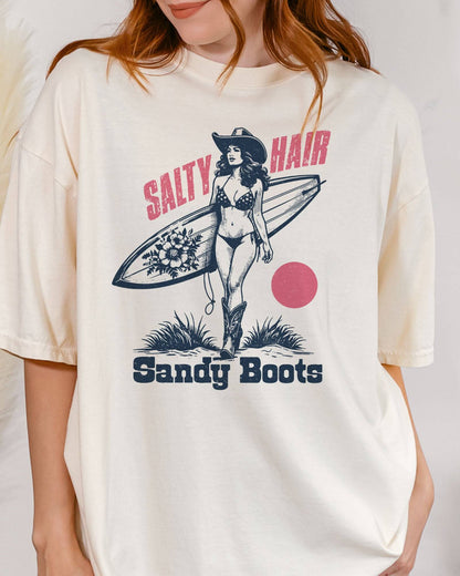 Close-up of woman wearing "Salty Hair Sandy Boots" shirt in white color with retro graphic of a cowgirl with a surfboard under her arm.