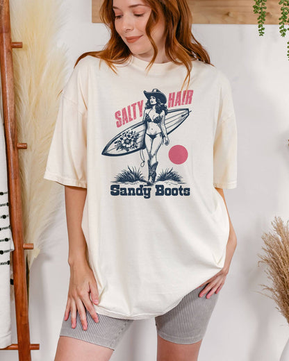Woman wearing "Salty Hair Sandy Boots" shirt in ivory color with retro graphic of a cowgirl with a surfboard under her arm.